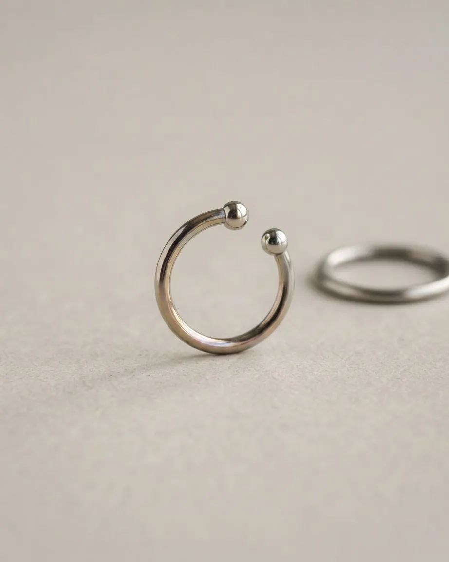 A silver circular septum jewelry ring with two ball ends rests on a pale studio surface.