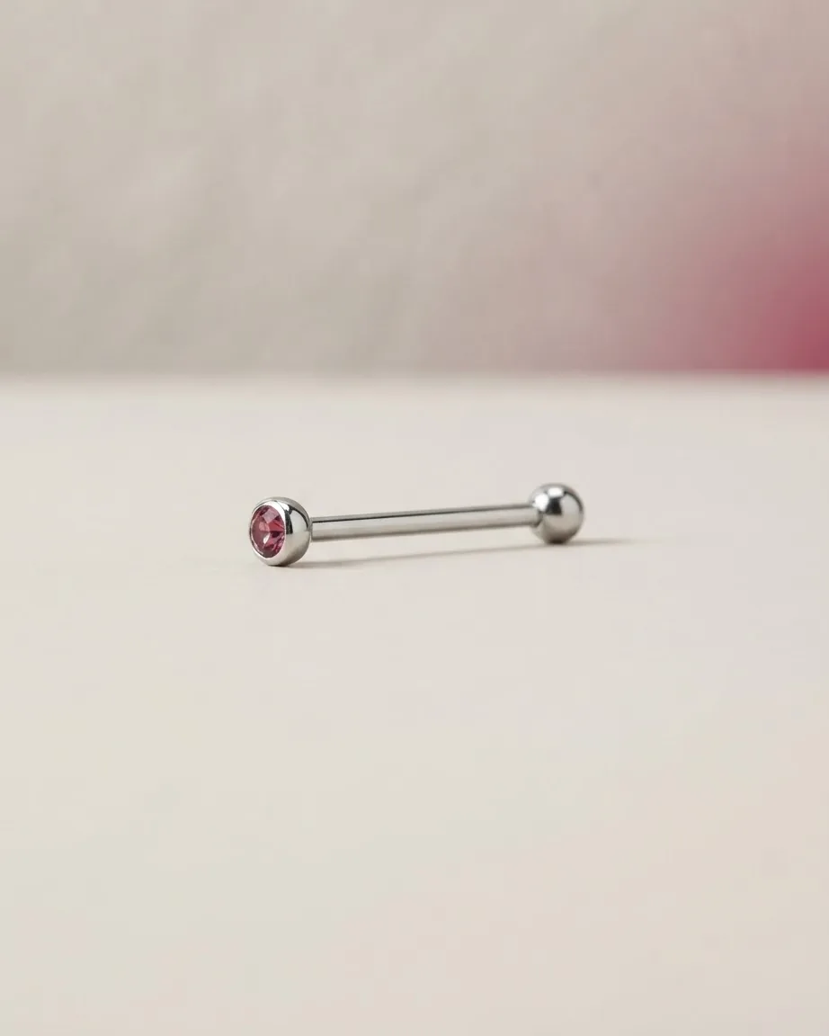 A silver straight barbell piercing with a pink gemstone on one end rests on a smooth cream-colored surface.