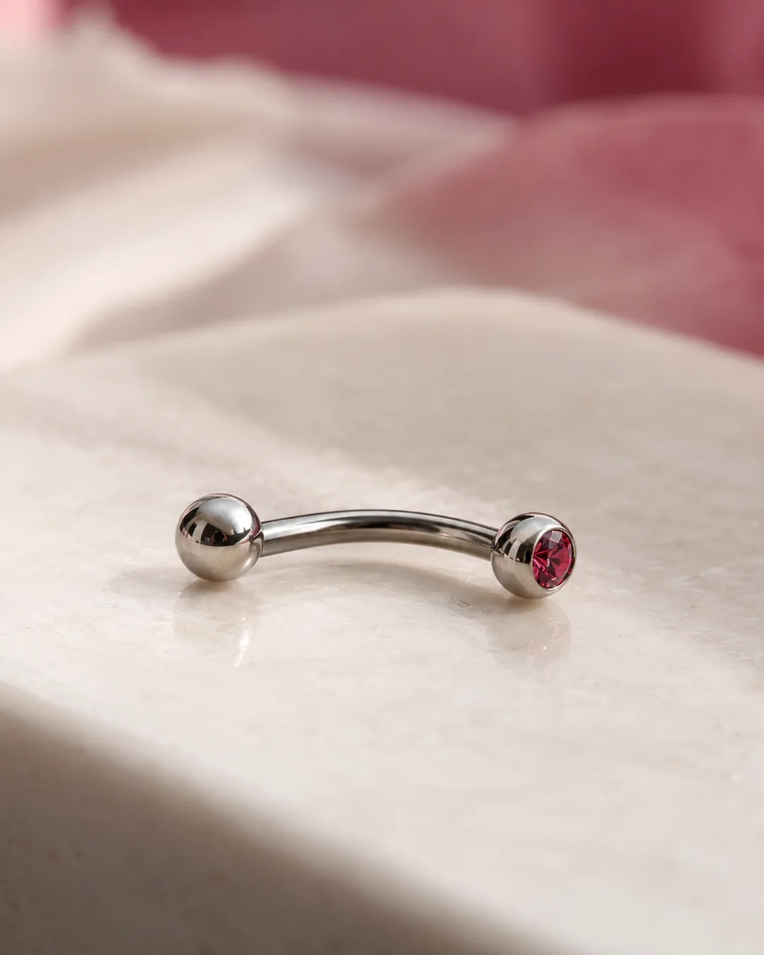 A polished silver curved barbell with one plain ball end and one pink gemstone end rests on a cream-colored surface.