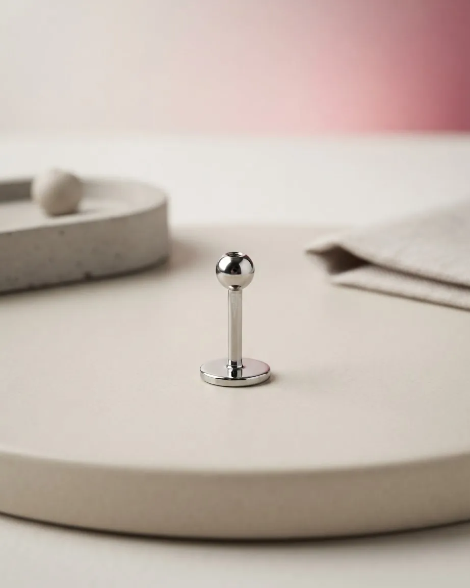 A polished silver flat-back labret stud stands upright on a beige surface beside a stone tray and folded fabric.