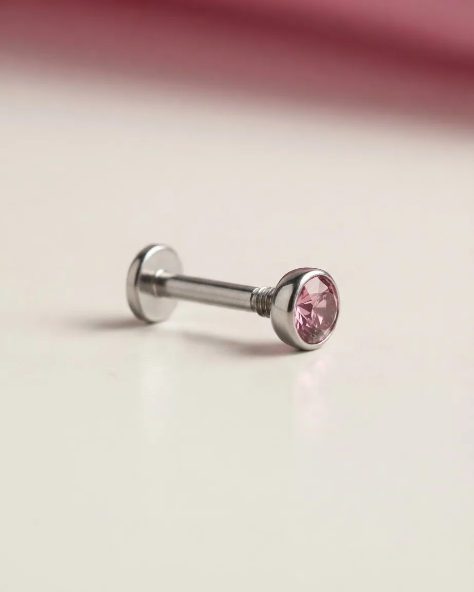 A silver flat-back labret stud with a round pink gemstone rests sideways on a smooth cream-colored surface.