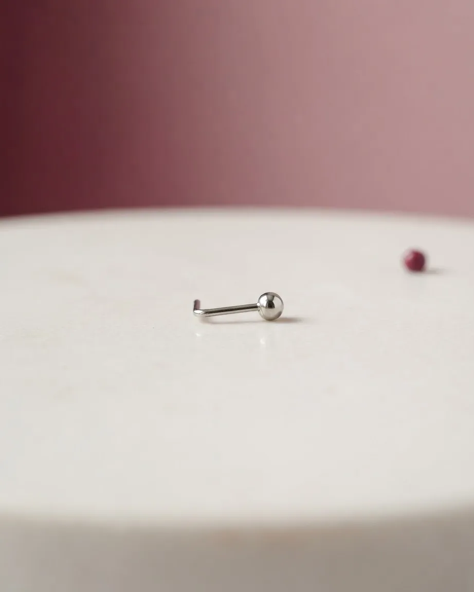 A small silver nostril post with a ball end rests on a pale surface against a blurred burgundy background.