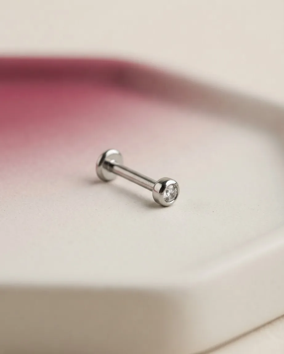 A small silver barbell piercing stud with a clear gemstone rests on a pale surface against a blurred pink background.