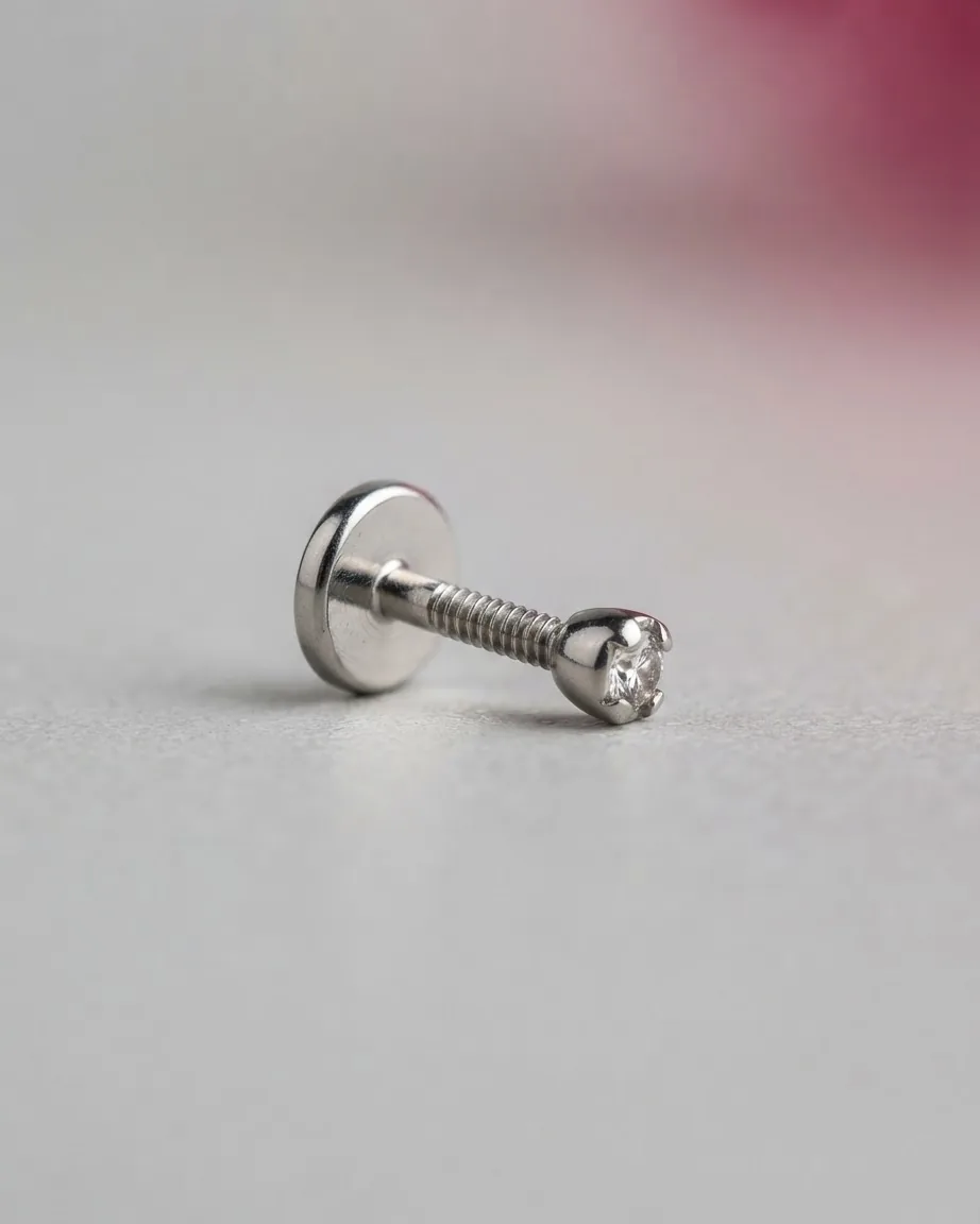 A small silver flat-back piercing stud with a clear gemstone rests sideways on a smooth gray surface.