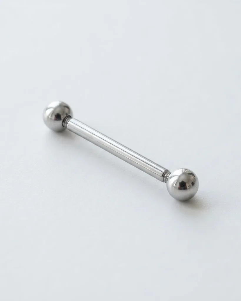 A polished silver barbell piercing with round metal balls at both ends rests diagonally on a pale gray surface.