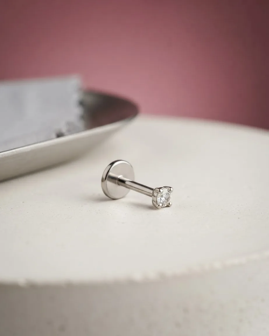 A silver flat-back labret stud with a clear gemstone rests on a cream surface beside a metal tray.